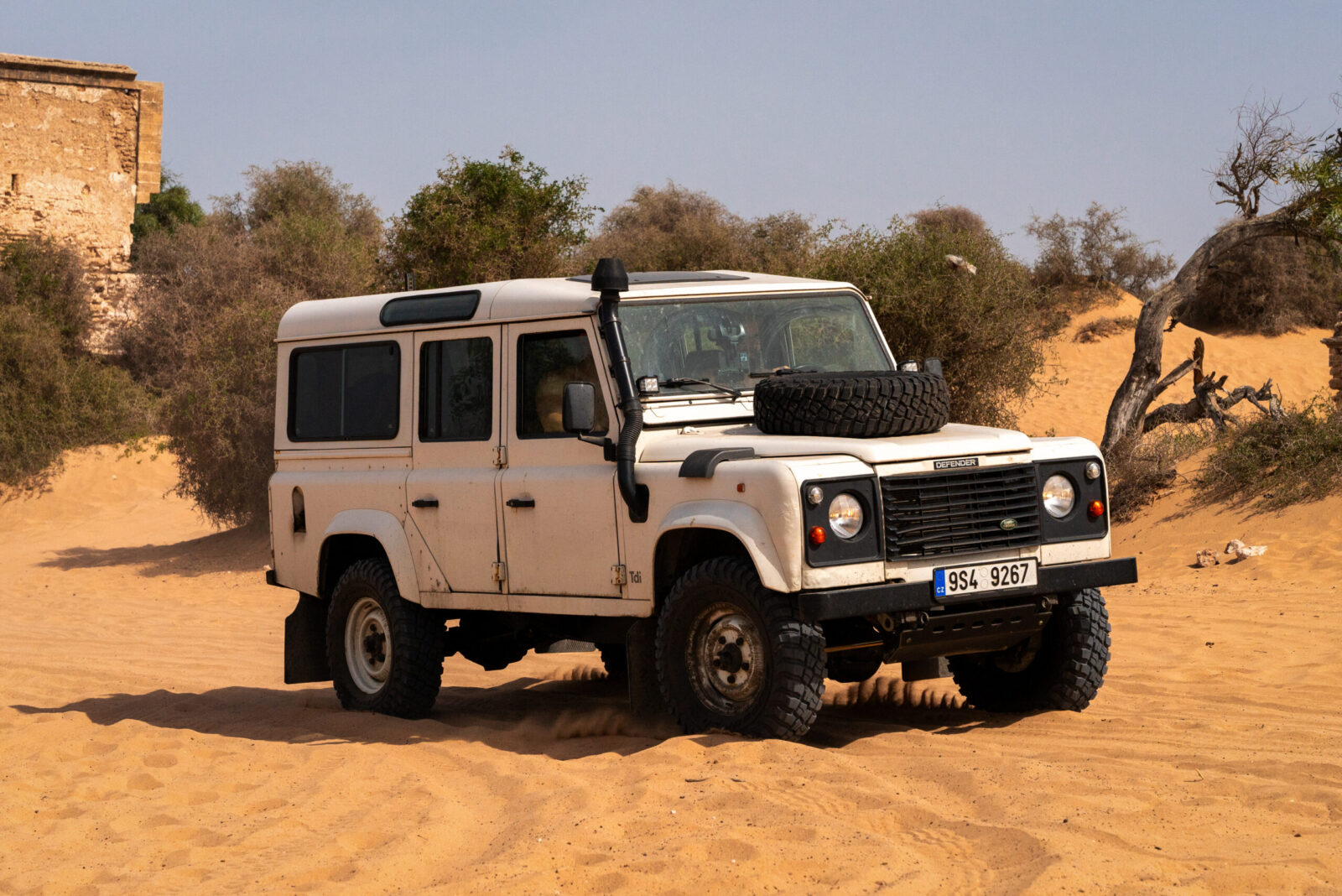 Bílý Land Rover Defender s rezervním kolem na kapotě u zříceniny paláce Dar Sultan v písečných dunách Maroka.