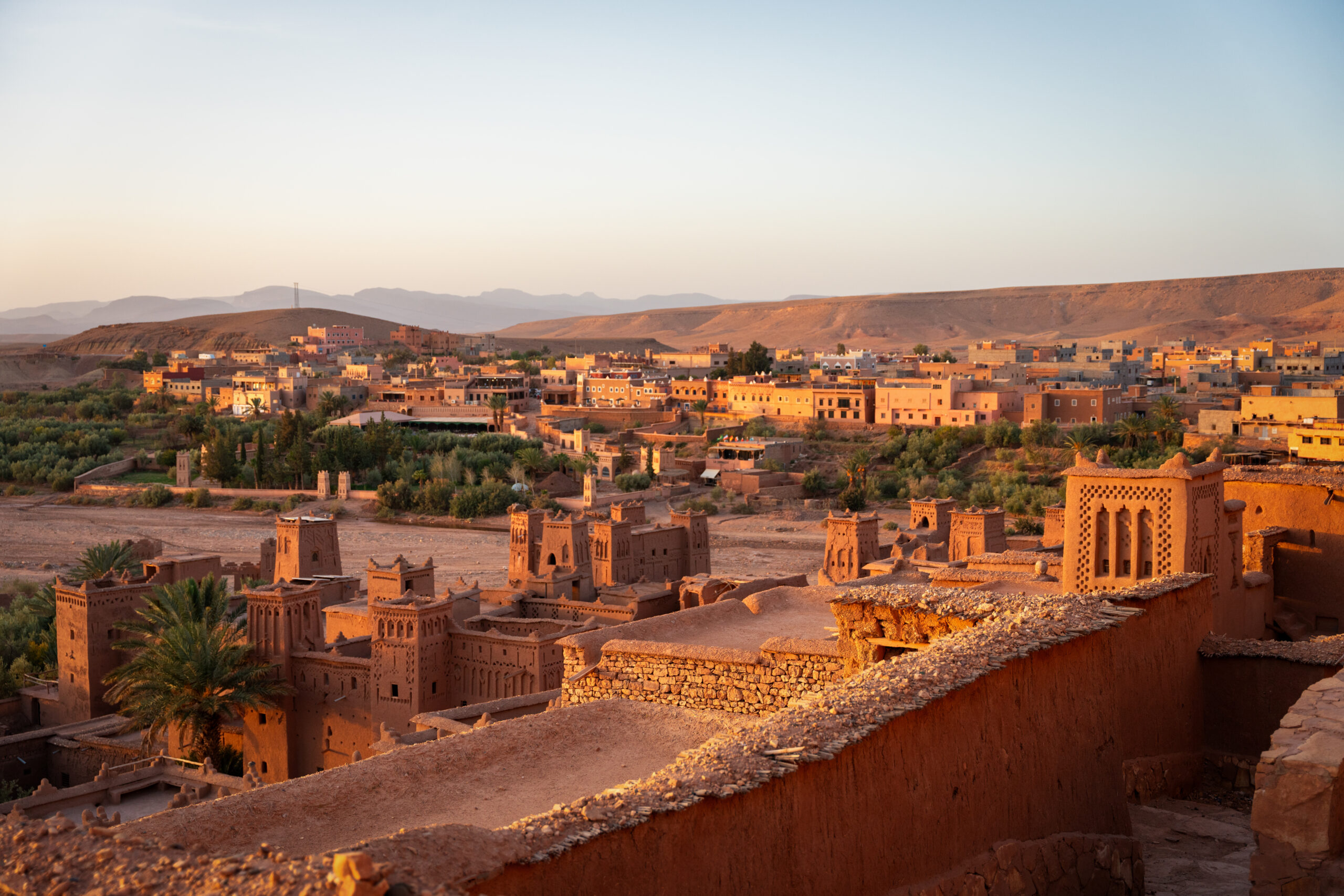 Pohled ze střechy ksar Aït Ben Haddou na věže z nepálené hlíny, palmovou oázu a novou část vesnice v Maroku při západu slunce.