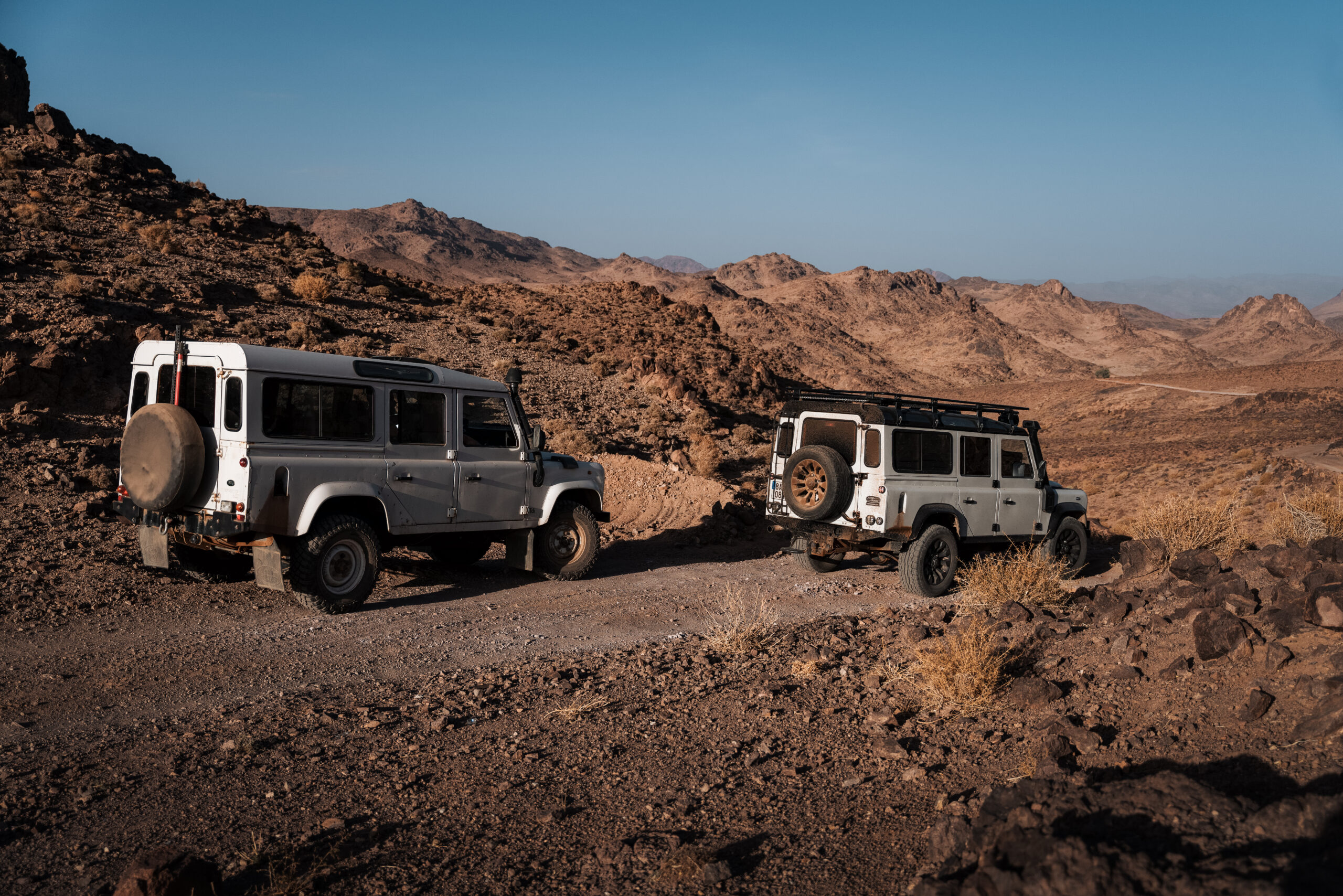 Dva terénní vozy Land Rover Defender na úzké kamenité cestě v pustém marockém pohoří.