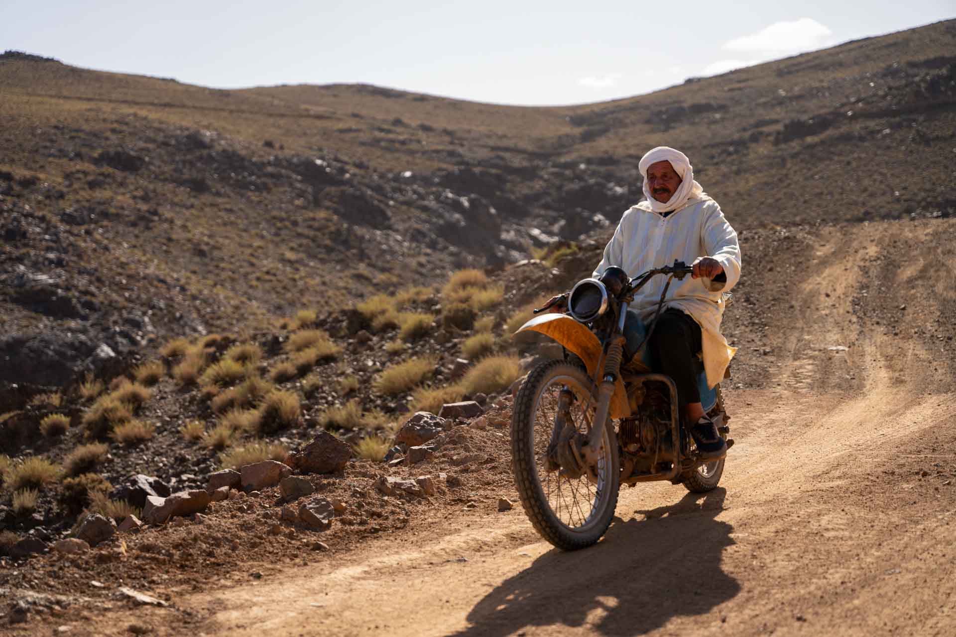Marocký muž v tradičním bílém oděvu a turbanu jede na motorce po prašné cestě v horách.