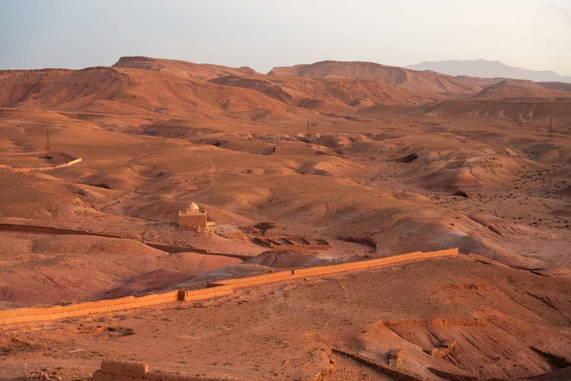 Pohled z kopce v Aït Ben Haddou na pouštní krajinu s obrannou zdí a malou hliněnou stavbou při západu slunce.