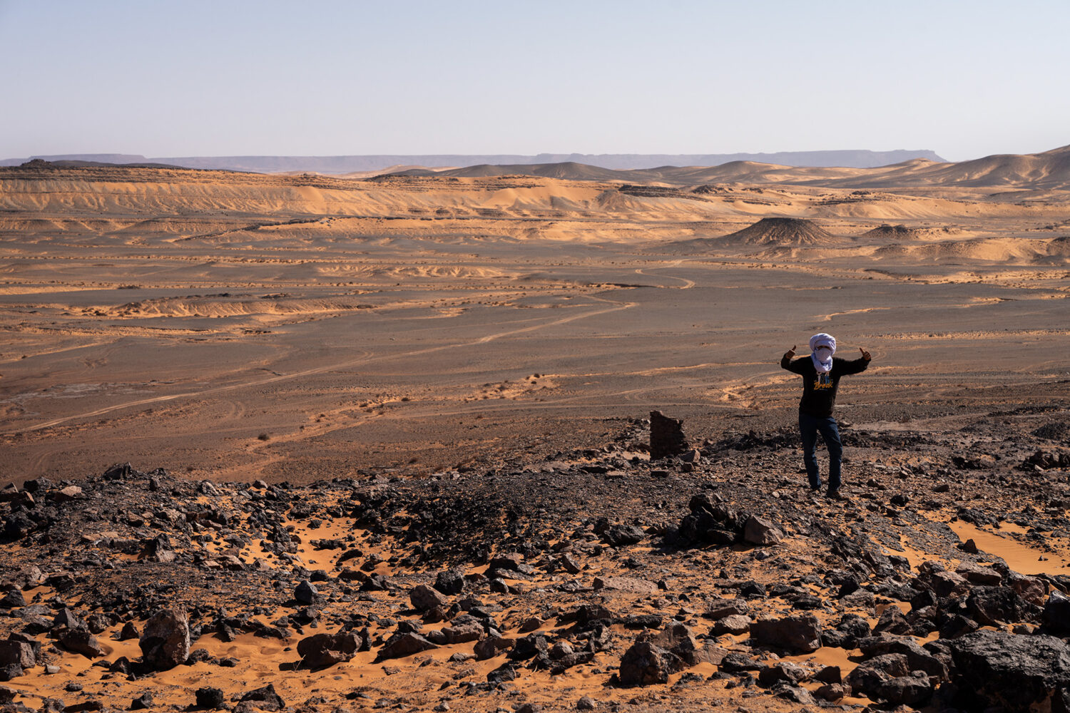 Muž v pouštním šátku na kamenitém kopci s výhledem na rozlehlé panorama pouště a dun u dolů M'Fis v oblasti Merzouga v Maroku, cestovatelská fotografie