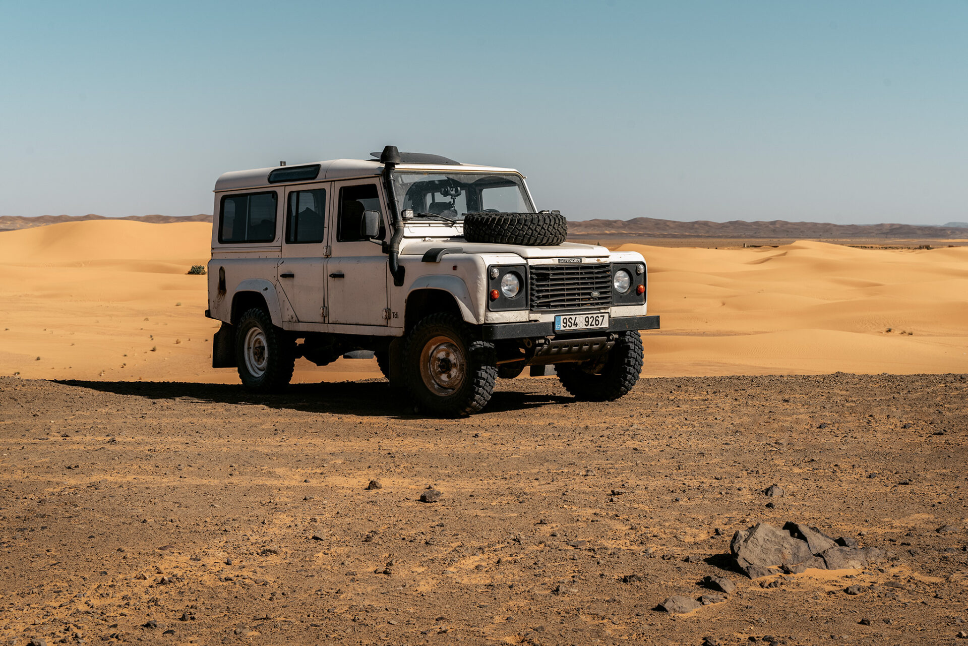 Expediční auto Land Rover Defender na kamenité poušti s výhledem na písečné duny Erg Chebbi v Merzouze v Maroku, off-road cestování